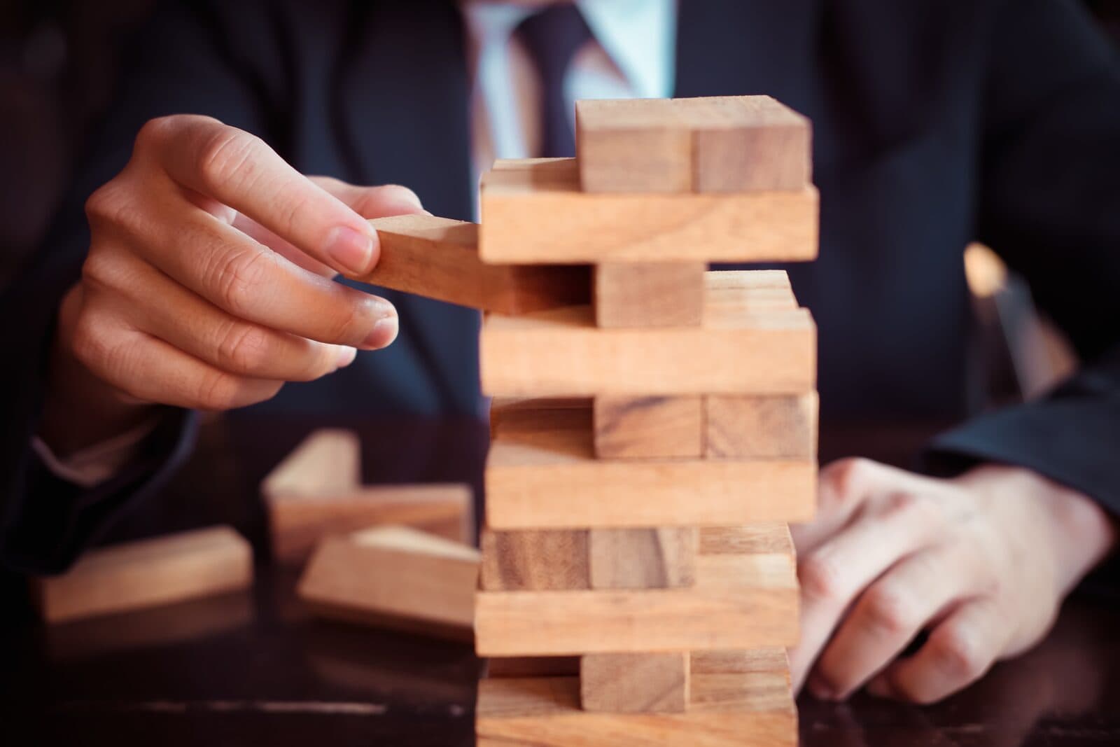 Business professional in a suit carefully removing a wooden block from a Jenga tower, representing the delicate balance and strategic decisions involved in business transitions and owner step-back